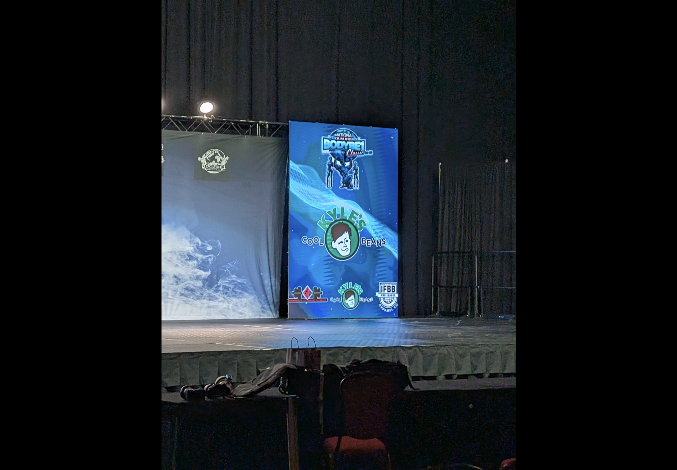 Stage with a large screen displaying Kyle’s Cool Beans and BodyBE1 logos and images, with a dark curtain background.