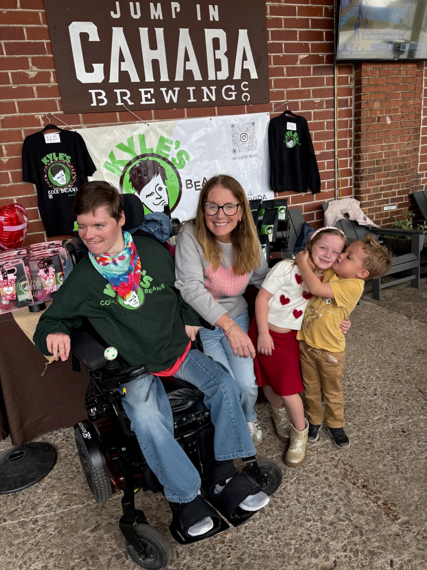 Kyle in a wheelchair with two children in front of a 'Cahaba Brewing' and Kyles Cool Beans sign and merchandise display.