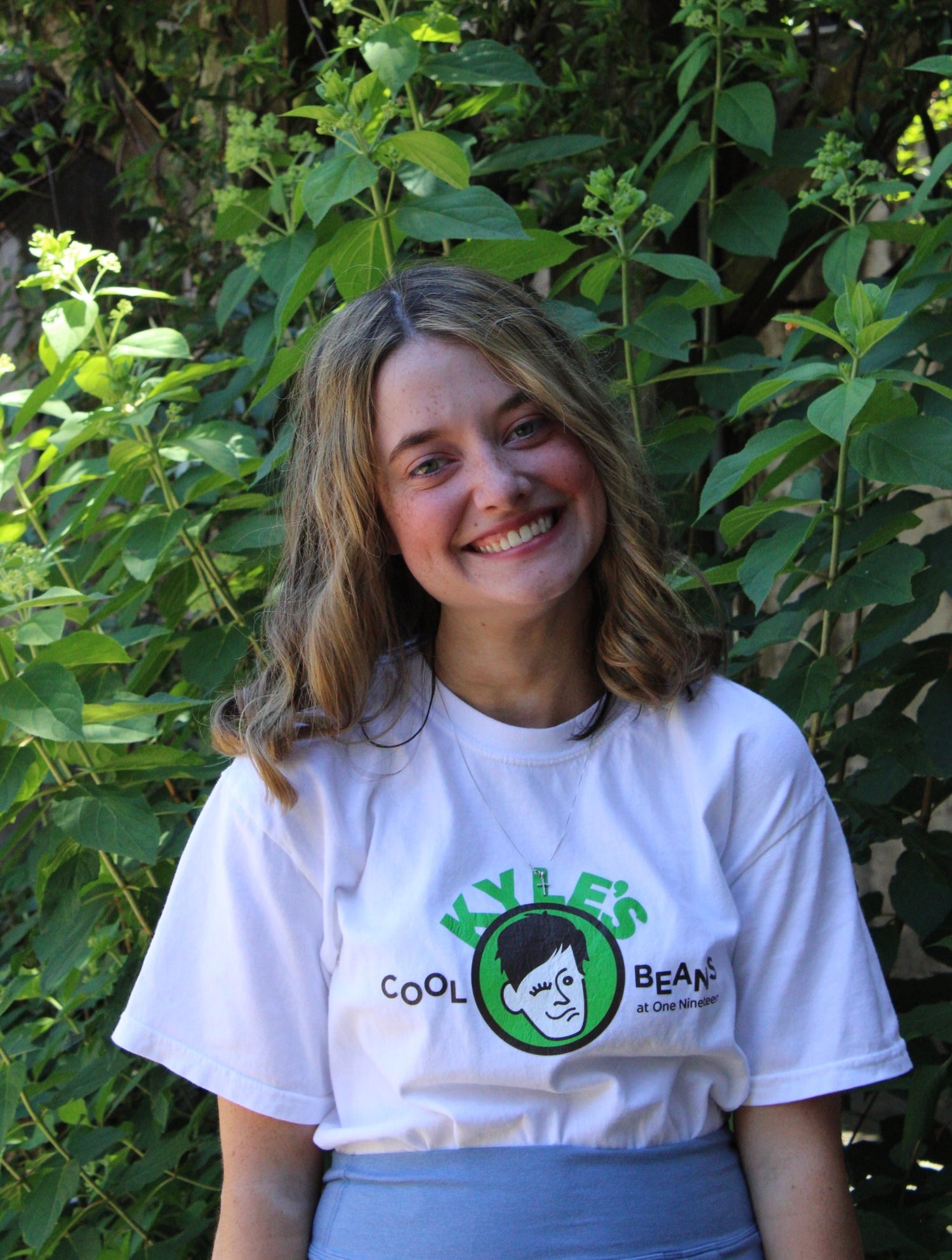Lizzie one of Kyle’s friends. Here she is pictured wearing a Kyle’s Cool Beans Shirt and smiling while standing in front of some leafy plants